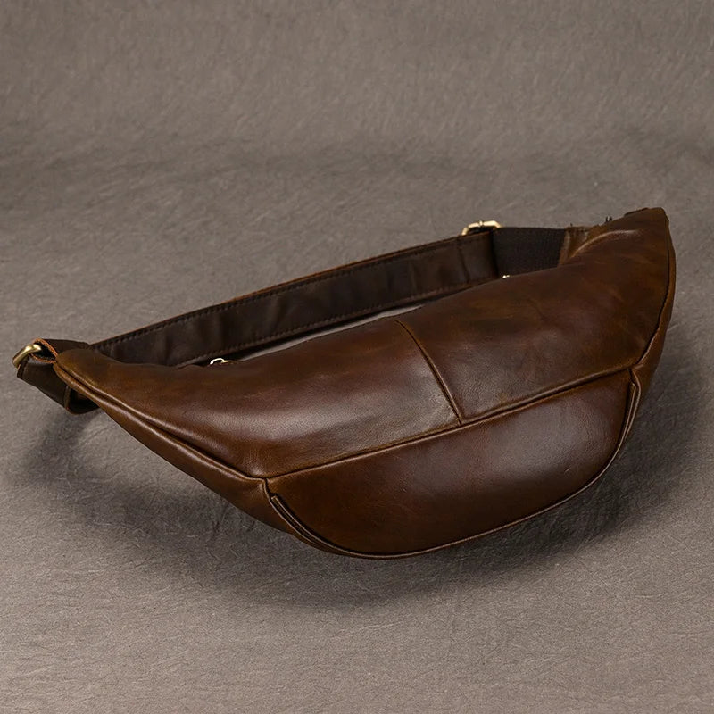 The Leather Belt Bag, crafted from genuine cowhide leather with a smooth finish and visible stitching, sits on a neutral, textured surface. The bag features a sleek, curved design and is equipped with an adjustable strap adorned with durable alloy hardware. Its interior is lined in black fabric.