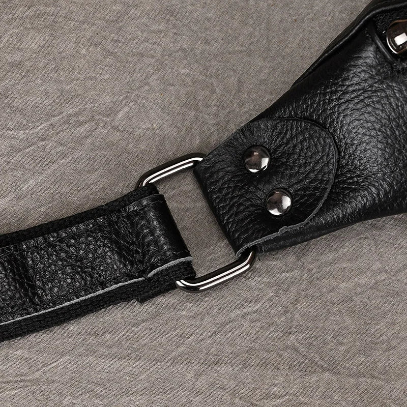 Close-up of a section of a black leather strap with a textured surface, connected by a polished silver rectangular metal ring. The strap, integral to the versatile design of the Black Leather Belt Bag, is secured with two silver rivets on a grey fabric background.