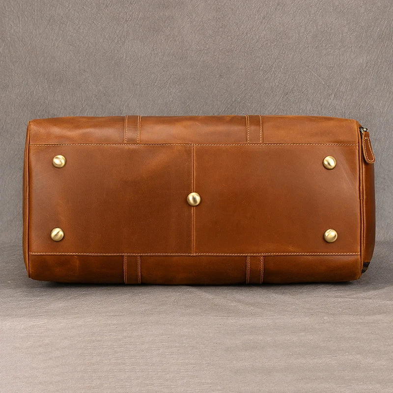 The Weekender Duffle 22 Leather Travel Bag, a large rectangular brown duffle, rests on a textured grey surface. This genuine cowhide leather bag features brass studs at its base for protection and has a smooth texture with visible stitching that enhances its sophisticated design.