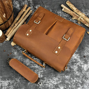 A Leather 14" Briefcase Shoulder Bag in brown is displayed on a gray textured surface. The bag features buckle straps and a handle, with its adjustable shoulder strap detached and lying beside the genuine cowhide piece. Wooden sticks and logs encircle the setup, enhancing the rustic ambiance.