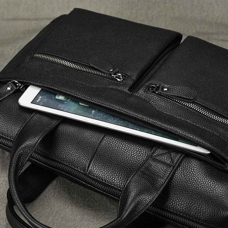 A 15" Black Briefcase, made of genuine leather and featuring two zippered pockets, is slightly open with a tablet partially sticking out from one of the pockets. The bag, which has an adjustable strap, rests on a gray fabric surface. The tablet's screen faces up, displaying part of the home screen.