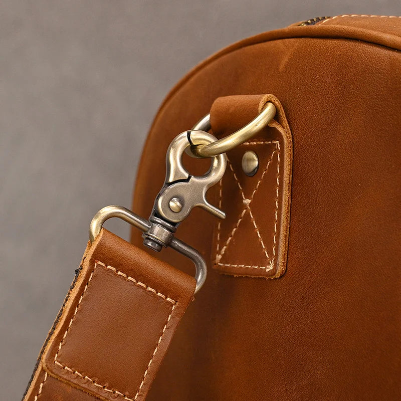 Close-up of a Weekender Duffle 22 Leather Travel Bag's strap and metal clasp detail. The strap is attached to a brass ring through a swivel hook clasp. The stitching on the genuine cowhide leather is visible, showcasing a neat, hand-crafted look. Perfect for a stylish travel companion, the background is a plain, light gray surface.