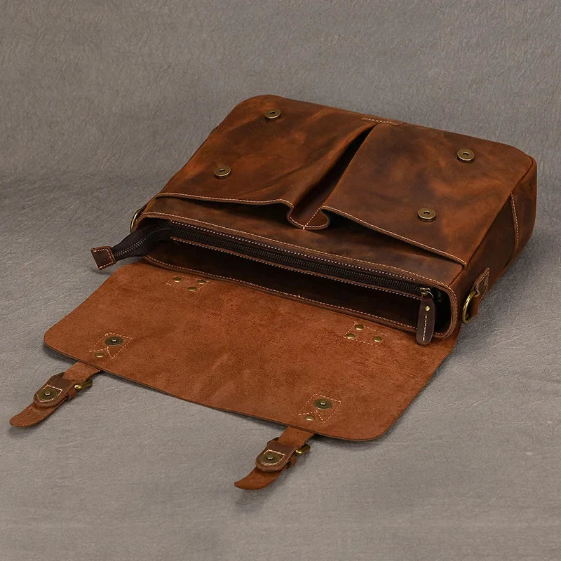 The Leather 14" Briefcase Shoulder Bag, crafted from genuine cowhide, is displayed open to reveal its spacious interior with a zipper compartment. This brown leather satchel features a flap opening and two adjustable straps with brass buckles and button snaps. The inner side of the flap is lined with suede, exuding a professional style against the gray background.