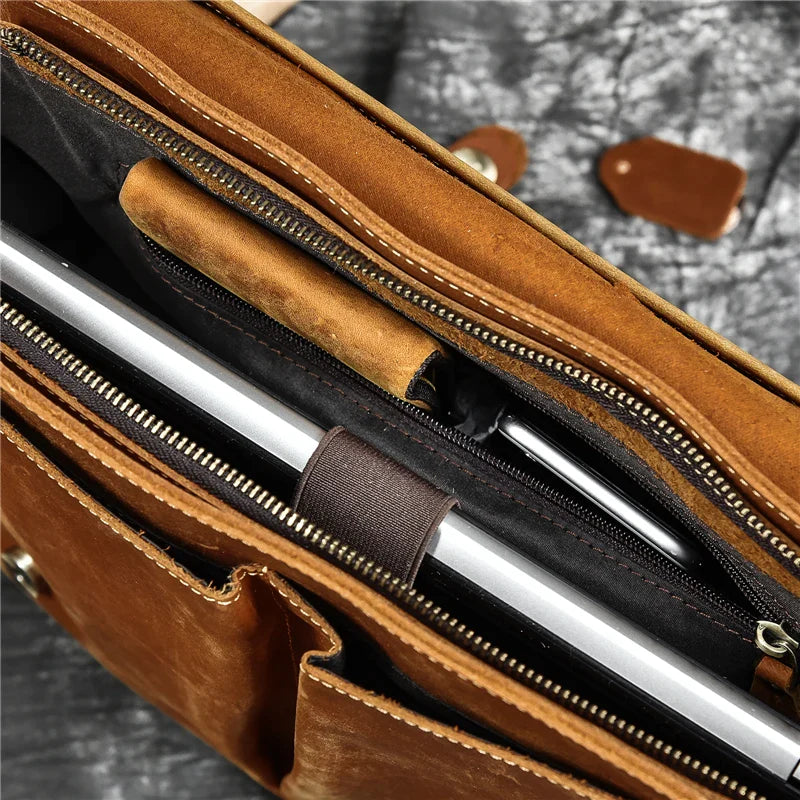 A top view of an open Leather 14" Briefcase Shoulder Bag shows multiple compartments crafted from genuine cowhide, one of which contains a partially visible laptop. The brown leather bag features zippers, a buckle strap, and additional pockets for organization. The background appears to be a gray textured surface—perfect for professional style.