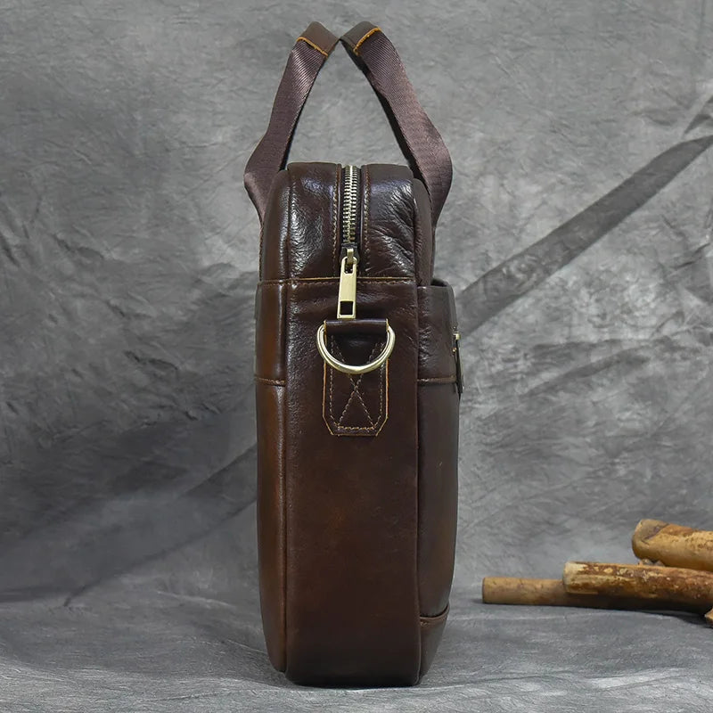 The 16" Brown Leather Briefcase stands upright on a textured gray background. The bag boasts a zipper closure, sturdy handles with dark brown detailing, and a metallic D-ring on its side, along with an adjustable strap. Some wooden props are partially visible in the background to the right.