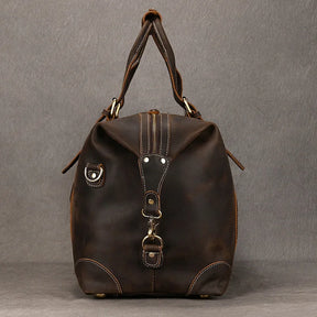 The Weekender 20 Leather Travel Duffle, a brown leather travel bag with reinforced stitching, is standing upright against a plain gray background. Crafted from genuine cowhide leather, the bag boasts multiple buckles and straps, metal accents, and includes a removable strap. The handles are neatly folded together at the top.