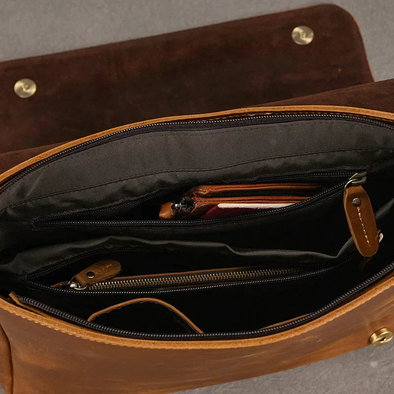 A top-down perspective of the open Vintage Leather Shoulder Bag reveals its multiple compartments. Inside, visible items include a red wallet, a zippered black pouch, and a tan cardholder. Crafted from genuine cow leather, the bag is lined with black fabric and secured by two brass buttons on the flap.