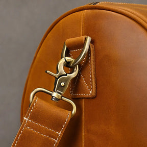 Close-up of a vintage Leather Travel Duffle in brown leather, featuring a metal clasp and strap. Visible stitching highlights expert craftsmanship. The focus on the attachment point emphasizes the bag's quality and design—ideal for stylish travel enthusiasts.