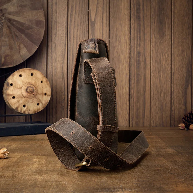 A Vintage Leather Shoulder Bag made from genuine cow leather rests on a wooden surface against a rustic wooden backdrop. The bag features a timeless style with an adjustable strap that has multiple holes for sizing. In the background, decorative elements, including a round wooden piece, add charm.