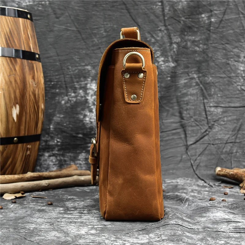 Side view of a Leather 14" Briefcase Shoulder Bag made from genuine brown cowhide leather placed against a dark textured background. A wooden barrel is partially visible on the left, and a few wooden sticks are scattered on the ground nearby. The briefcase, resembling a professional-style satchel, has metal hardware and appears to be in new condition.