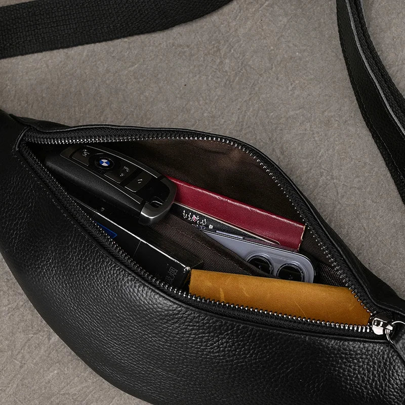 The Black Leather Belt Bag, crafted from premium leather, features a versatile design with a partially open zipper revealing car keys, a folded wallet, and cards. Its sleek strap lies elegantly on the grey surface.