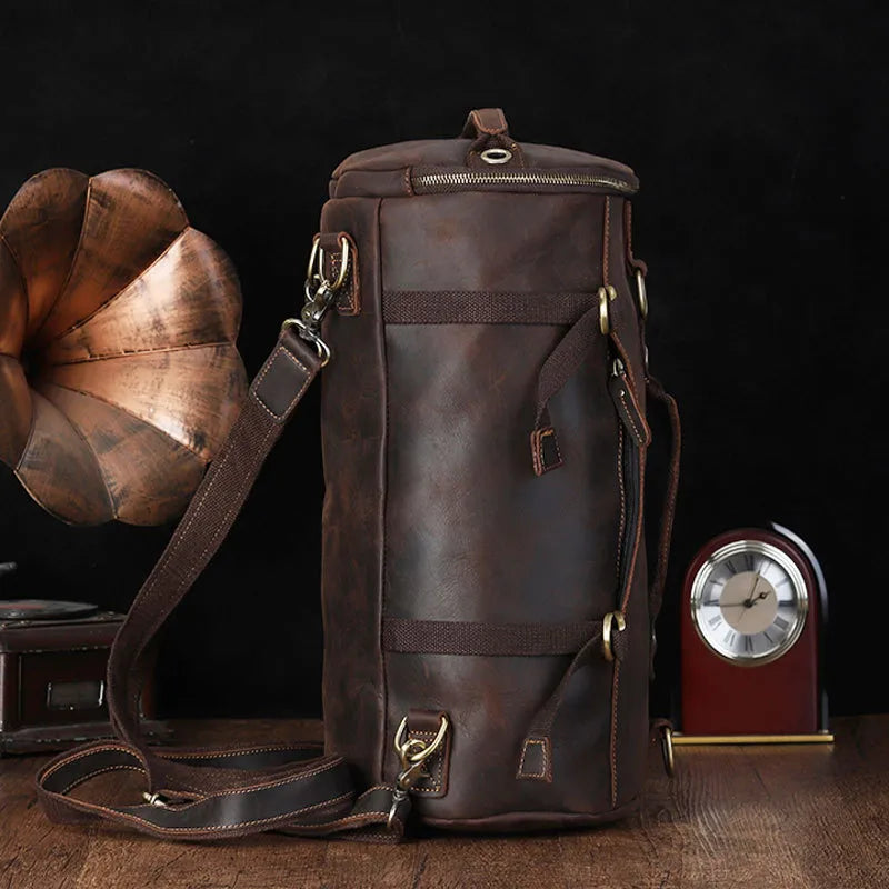 A Vintage Leather Travel Backpack crafted from genuine cowhide leather with brass fixtures is displayed on a wooden surface. To the left, part of a gramophone is visible. A red and silver clock sits to the right, set against a dark background, perfect for the stylish traveler.