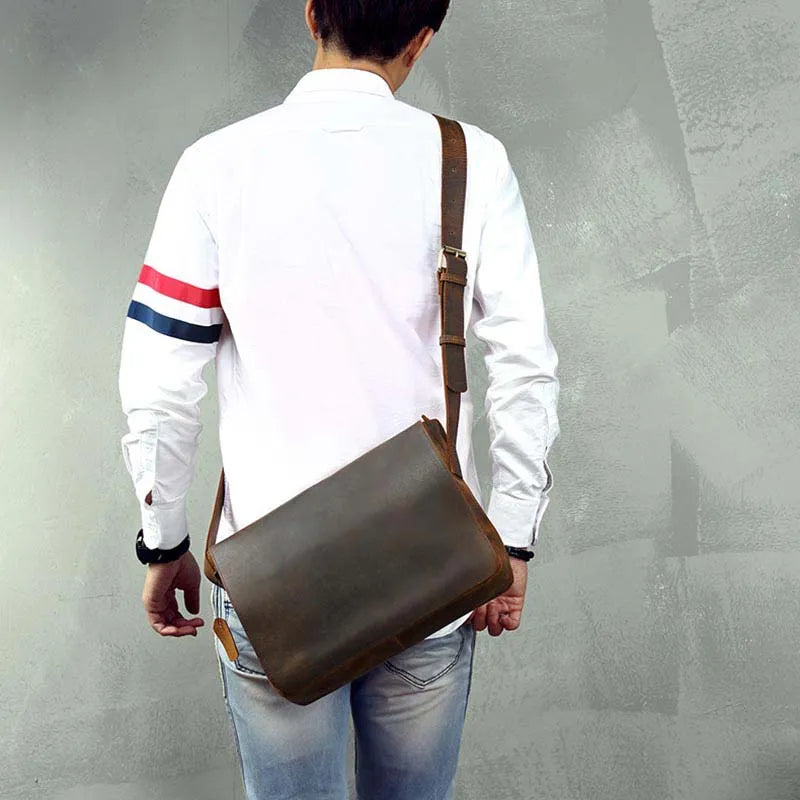 A person stands with their back to the camera, wearing a white shirt with blue, white, and red striped sleeves paired with light blue jeans. They are holding the Vintage Leather Shoulder Bag against a subtly textured gray background.