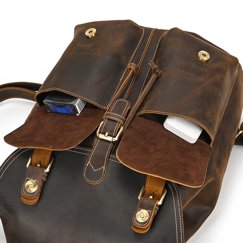 The Genuine Leather Vintage Backpack for men in brown showcases an open flap that reveals two pockets: one designed to hold a pack of cigarettes and the other for a sleek white smartphone. The casual style is enhanced by brass buckle details and a belt strap closure, making it ideal for everyday adventures.