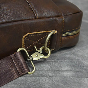 Close-up image of the 16" Brown Leather Briefcase featuring a genuine leather finish, metal clasp and buckle attaching an adjustable dark brown strap. The stitch work and texture of the leather are visible alongside a zipper on one side of the bag. The background is a gray, textured surface.