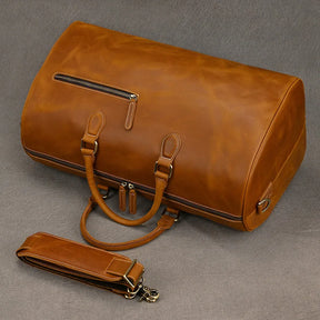 The Leather Travel Duffle, a large vintage-style brown bag, is crafted from genuine leather with a smooth finish. It includes sturdy handles, an adjustable shoulder strap, and a side zippered pocket. The bag is displayed against a plain gray background.