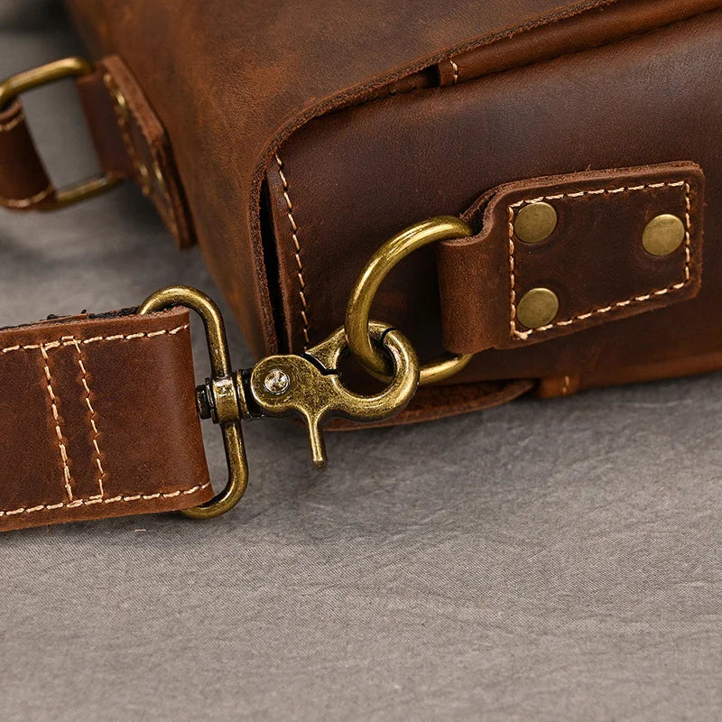 Close-up of the Leather 14" Briefcase Shoulder Bag showcases its vintage brown leather with brass hardware. The image highlights a sturdy clasp attaching a strap to the bag. The stitching and detailing on the genuine cowhide, as well as the riveted brass hardware, are prominently displayed, enhancing its professional style.