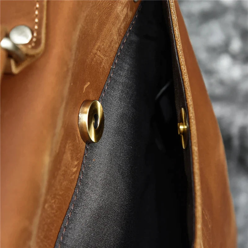 Close-up of an open Leather 14" Briefcase Shoulder Bag made from genuine cowhide leather, featuring a gold snap button. The bag's interior is lined with dark fabric, with detailed stitching visible along the edge. The background is blurred and appears to be gray, enhancing its professional style.