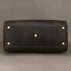 The bottom view of the Leather Travel Duffle, a vintage brown leather bag featuring gold studs, a central gold rivet, and visible stitching with a slightly distressed finish, is set against a gray background.