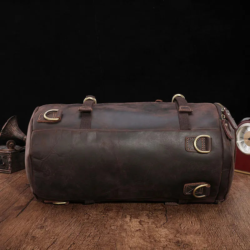 A Vintage Leather Travel Backpack in a dark brown, made from genuine cowhide leather and adorned with brass buckles, rests elegantly on a wooden surface. Ideal for the stylish traveler, it features a cylindrical design with multiple strap attachments. In the background, a gramophone and clock add to the timeless scene.