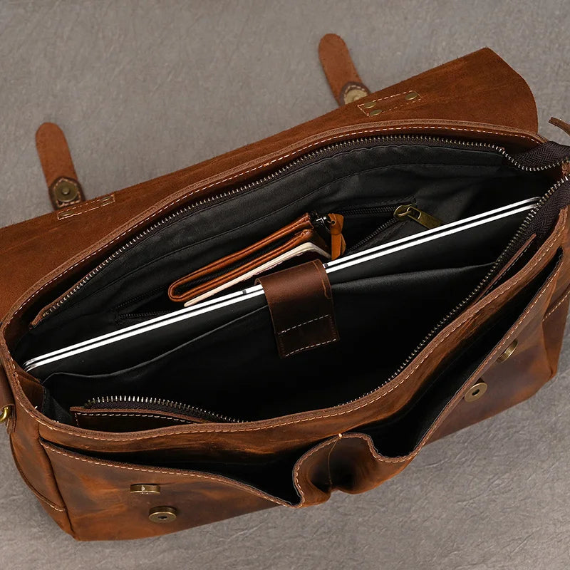 Top view of an open Leather 14" Briefcase Shoulder Bag revealing its contents. Inside, there is a laptop, a black pouch, and a small brown case. The briefcase has multiple compartments and pockets, featuring a brass snap closure. The genuine cowhide exhibits quality craftsmanship against the textured grey background.