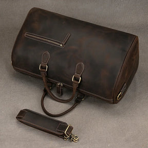 The Leather Travel Duffle, a spacious vintage dark brown leather bag with sturdy handles and a detachable shoulder strap, rests on a gray surface. Made from genuine leather, it includes a small zippered side pocket and brass hardware accents—a perfect choice for timeless travel.