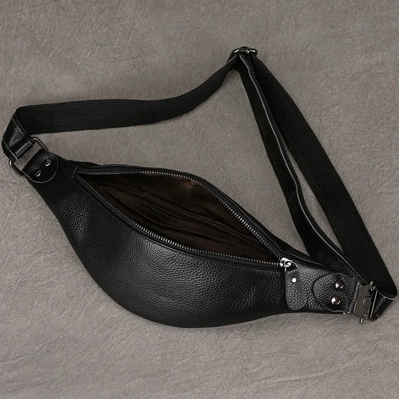 A Black Leather Belt Bag, crafted from premium leather with silver hardware, lies flat on a neutral gray surface. This black fanny pack features an adjustable strap and a partially open zipper, revealing the inner lining—exemplifying its versatile design.