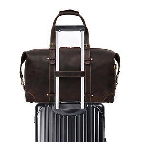 The Weekender 20 Leather Travel Duffle, crafted from genuine cowhide leather in a dark brown shade, is securely perched on top of a black hard-shell rolling suitcase. The duffle's handles are fitted over the suitcase handle, showcasing a convenient travel setup against a plain white background.