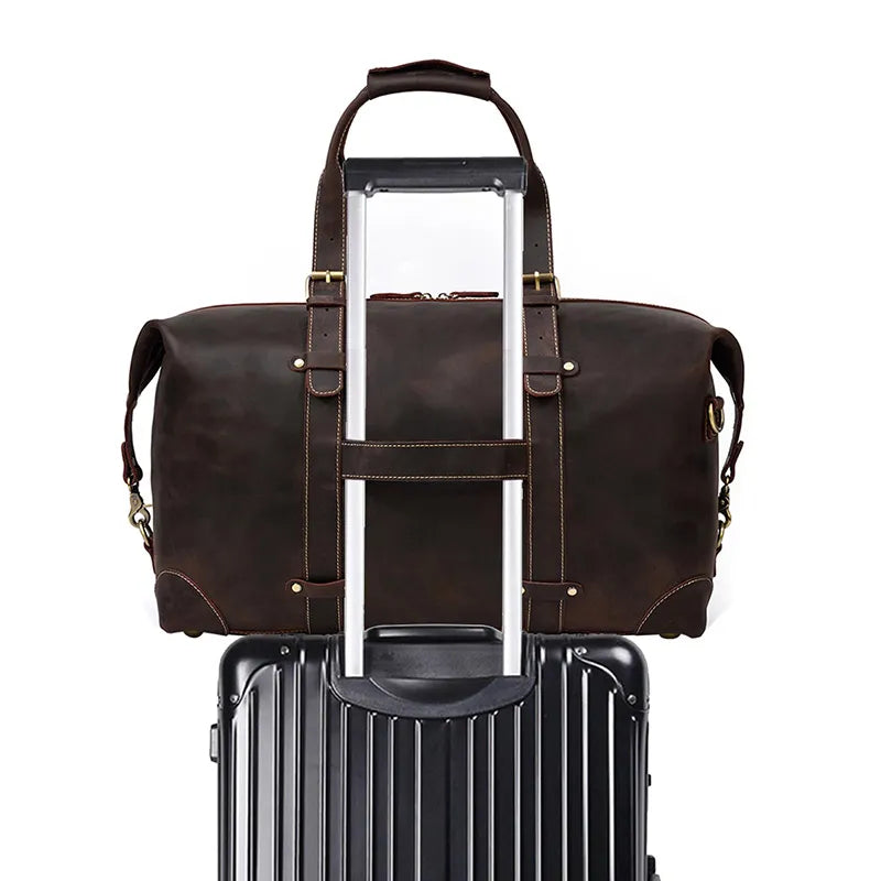 The Weekender 20 Leather Travel Duffle, crafted from genuine cowhide leather in a dark brown shade, is securely perched on top of a black hard-shell rolling suitcase. The duffle's handles are fitted over the suitcase handle, showcasing a convenient travel setup against a plain white background.