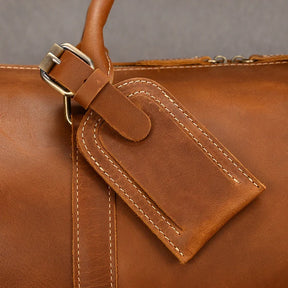 Close-up of the Weekender Duffle 22 Leather Travel Bag in brown leather, showcasing a stitched luggage tag attached with a metal buckle. The tag is rectangular with rounded edges and has a strap looped through the buckle for secure attachment. The Genuine Cowhide Leather bag is set against a neutral grey background.