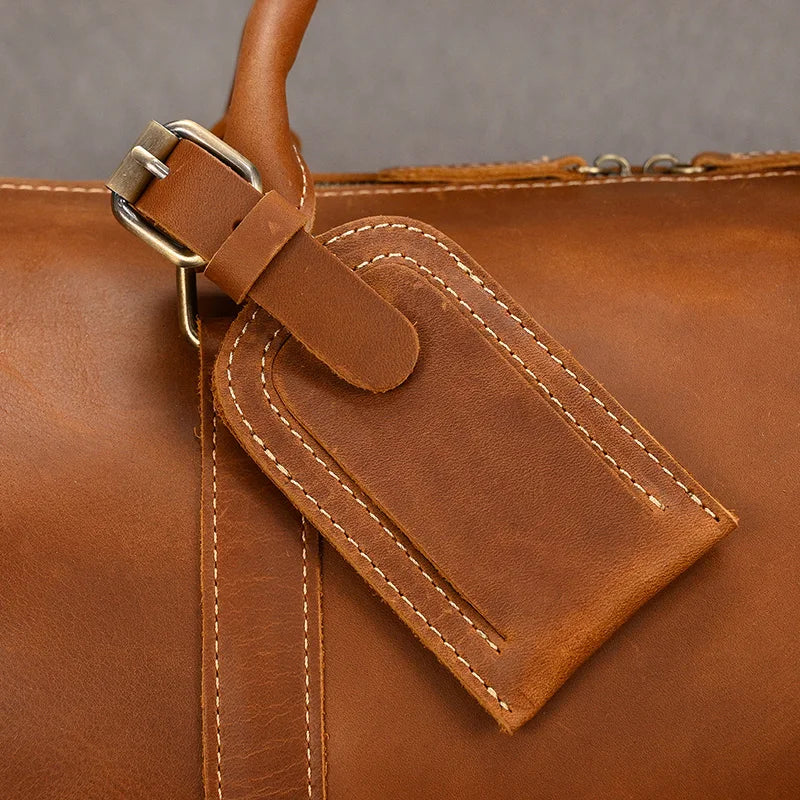 Close-up of the Weekender Duffle 22 Leather Travel Bag in brown leather, showcasing a stitched luggage tag attached with a metal buckle. The tag is rectangular with rounded edges and has a strap looped through the buckle for secure attachment. The Genuine Cowhide Leather bag is set against a neutral grey background.