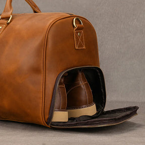 The Weekender Duffle 22 Leather Travel Bag, crafted from genuine cowhide leather and adorned with golden hardware, is partially open to reveal a pair of brown leather shoes tucked inside a side compartment. Its sturdy design is showcased against a plain, grey background.