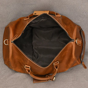 A top-down view of an open, empty Weekender Duffle 22 Leather Travel Bag made from genuine cowhide leather reveals a black interior. The bag showcases silver metal hardware, including a zipper and rings, and is placed on a grey surface. Two handles and a detachable shoulder strap are also visible.