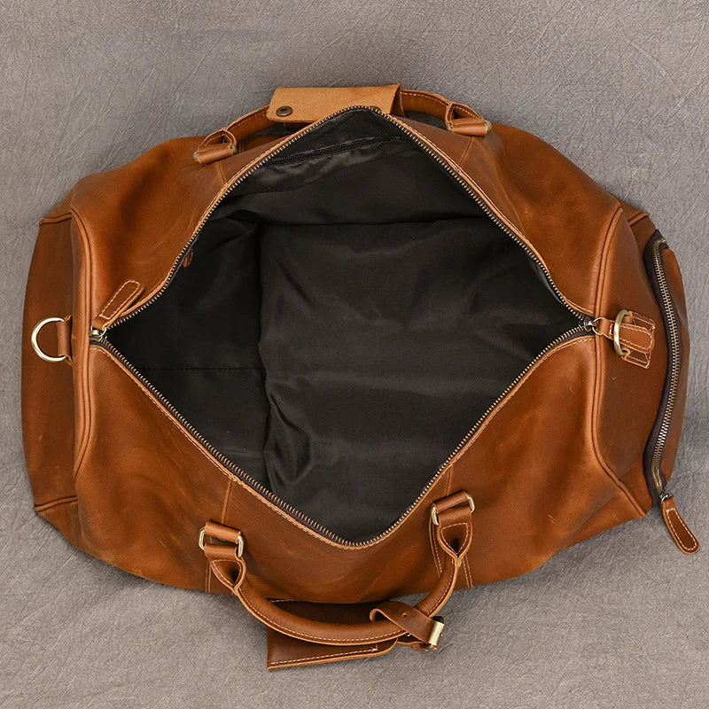 A top-down view of an open, empty Weekender Duffle 22 Leather Travel Bag made from genuine cowhide leather reveals a black interior. The bag showcases silver metal hardware, including a zipper and rings, and is placed on a grey surface. Two handles and a detachable shoulder strap are also visible.