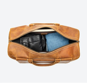 A top view of the Vintage Genuine Leather Travel Duffle Bag showcases its spacious interior, revealing a folded black garment and blue jeans. The premium travel bag, made from genuine cow leather, is set against a plain, light background.