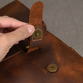 A hand is seen fastening a magnetic buckle on the Leather 14" Briefcase Shoulder Bag. The strap with a metal snap button is being secured to its corresponding clasp on the bag. The genuine cowhide leather has a rich, vintage texture. The focus is on the fastening mechanism, exuding a professional style.