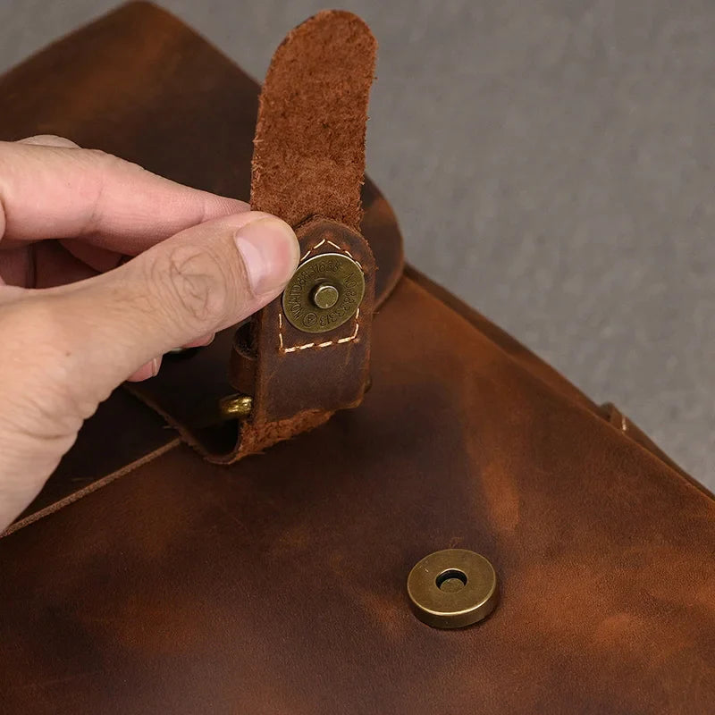 A close-up image of a hand unfastening the Leather 14" Briefcase Shoulder Bag, crafted from brown genuine cowhide leather with a brass magnetic clasp. The leather appears worn, complemented by a plain, neutral background.