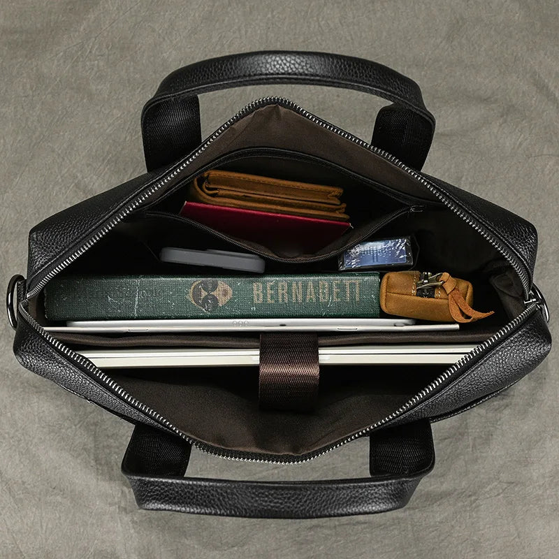 A 15" Black Briefcase in black leather is open, revealing its contents. Inside are a green book titled "BERNADETT," a brown wallet, a folded white paper, a pen, a small notebook, a smartphone, and another small item in a leather case. The bag features black handles, an adjustable strap, and a brown interior.