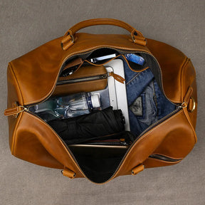 The Leather Travel Duffle, a brown genuine leather bag with a vintage zippered design and handles, is open displaying a clear water bottle, black umbrella, blue jeans and shirt, along with a digital device like a tablet or laptop.