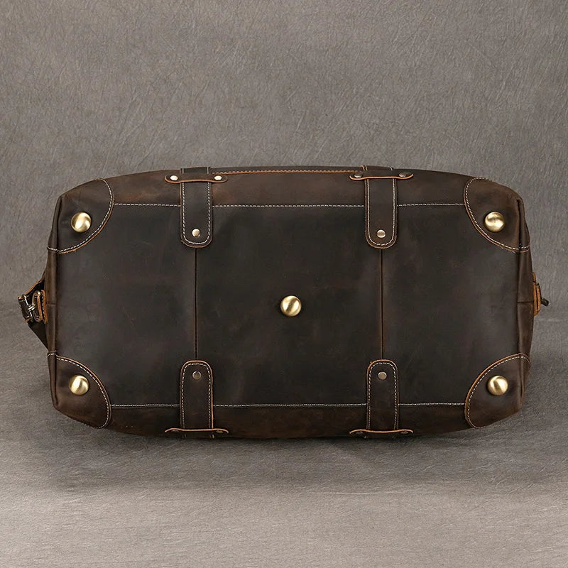 The Weekender 20 Leather Travel Duffle is displayed lying on its side, highlighting the bottom of the bag. It features metal studs on the corners and base, reinforced stitching, and a rich, dark finish in genuine cowhide leather. The background is a plain, textured gray surface.