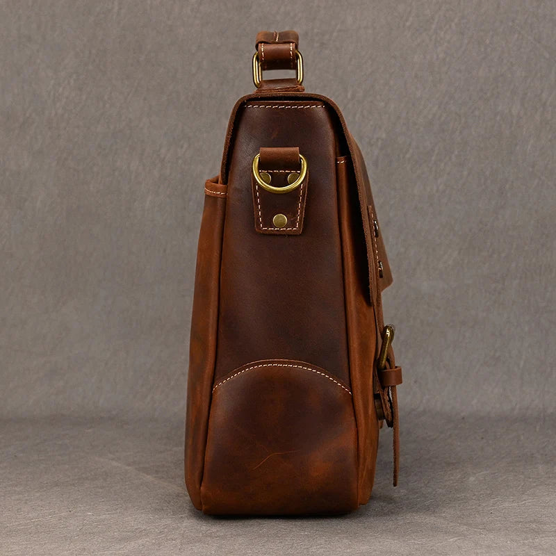 Side view of the Leather 14" Briefcase Shoulder Bag in brown, featuring a brass D-ring and clasp, detailed stitching, and a shoulder strap attachment at the top. The surface showcases genuine cowhide with a slightly worn texture, enhancing its professional style. The background is plain grey.