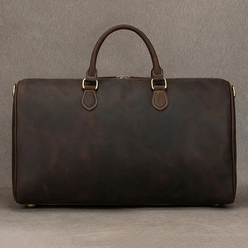 The Leather Travel Duffle is a vintage dark brown bag with sturdy handles and gold-tone hardware, set against a neutral background to emphasize its smooth texture and elegant design crafted from genuine leather.