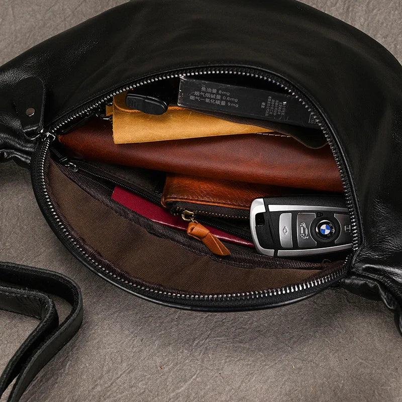 A black Leather Belt Bag, crafted from genuine cowhide leather, lies open, revealing its contents: a brown wallet, keys with a BMW key fob, a small black case, a yellow item (possibly a notebook or another small bag), and other miscellaneous items. The interior lining is brown and features durable alloy hardware.