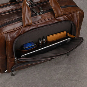 A brown leather 17" Business Travel Briefcase with a front pocket containing various items, including a tablet, two pens, a pack of blue tissues, and a brown leather wallet. Ideal for business travel, this genuine leather briefcase sits elegantly on a grey surface.