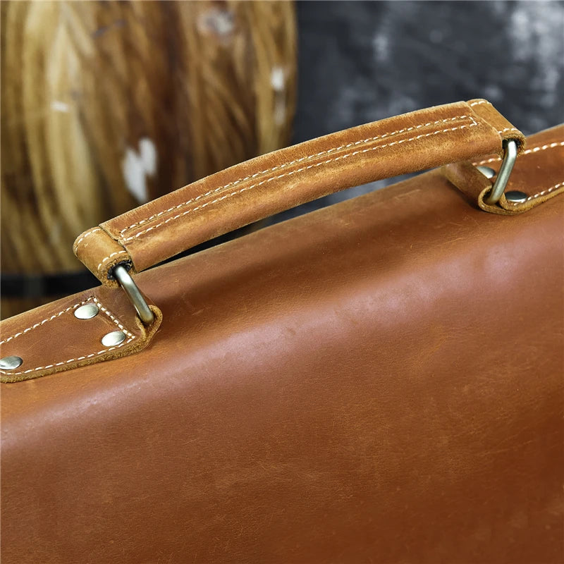 Close-up of the Leather 14" Briefcase Shoulder Bag's handle. The handle is secured with metal rings and rivets, and the genuine cowhide leather shows subtle texture and stitch details. There's a blurred, wooden background in muted tones, adding contrast to the leather's rich color and professional style.