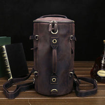 A versatile Vintage Leather Travel Backpack made from genuine cowhide leather rests on a wooden surface, accompanied by two books and a bottle of Hennessy Cognac against a dark background. Perfect for the stylish traveler, it boasts multiple straps, metal rings, a top handle, and adjustable shoulder straps.