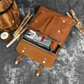 The Leather 14" Briefcase Shoulder Bag, crafted from genuine brown cowhide, is open, showcasing its contents: a laptop, a small book, and a pink smartphone. The briefcase rests on a dark textured surface surrounded by wooden logs and a rustic wooden barrel, radiating professional style.