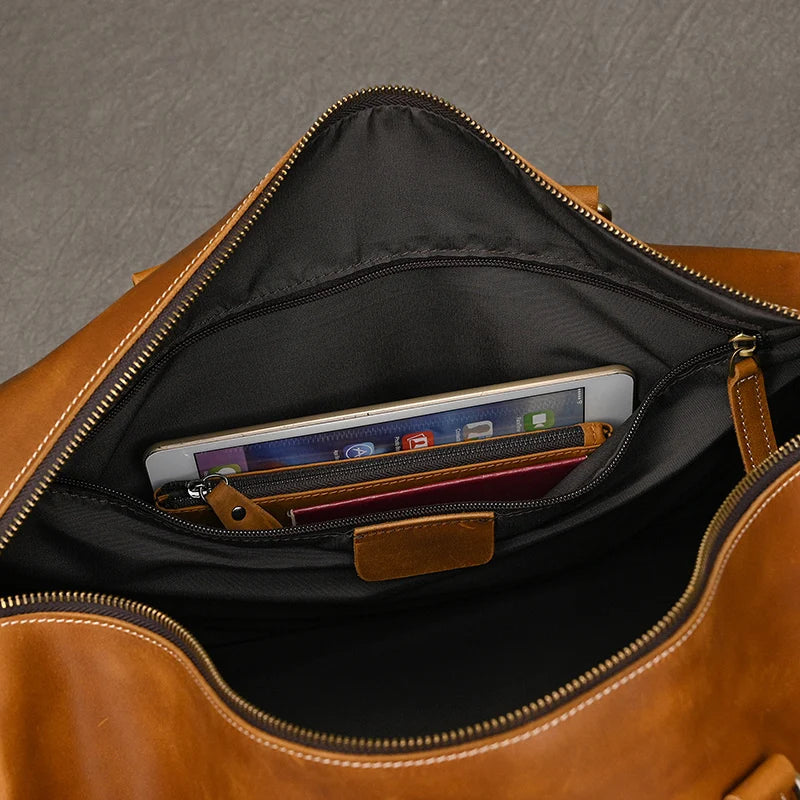 The Leather Travel Duffle unveils its essentials: a tablet, red wallet, and pen in the inner pocket. Unzipped to reveal its spacious black interior, this genuine leather travel companion rests elegantly against a dark surface.