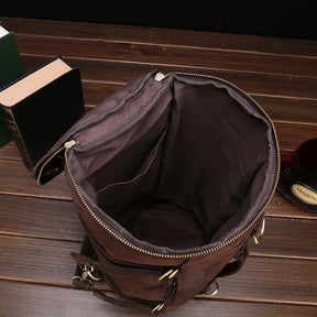 A Vintage Leather Travel Backpack, made from genuine cowhide leather, rests open on a wooden table. The versatile bag reveals a spacious interior with pockets. Nearby, there is a closed hardcover book and a partially visible bottle.