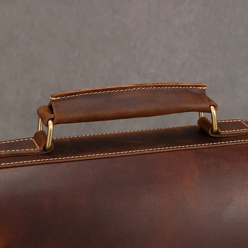 Close-up image of a brown leather handle on the Vintage Leather Briefcase. The handle is stitched with tan thread and attached to the bag with brass-colored metal rings. The background is a plain gray surface, highlighting the professional image of the accessory.